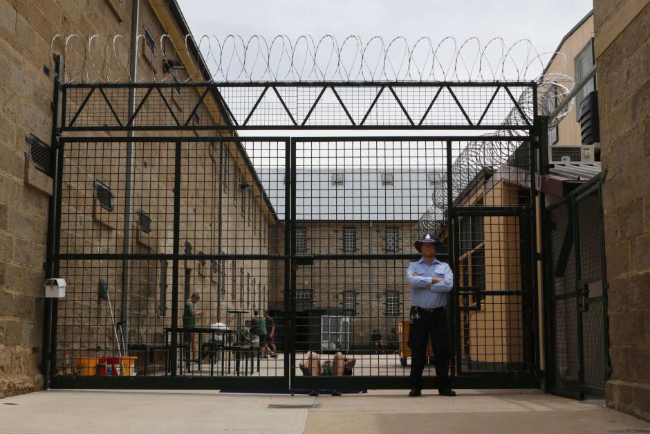 A guard stands in front of a razor wire fence behind which inmates can be seen talking and exercising.