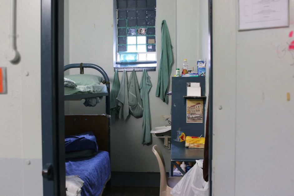 Through the door of a jail cell, a double bunk and a table and shelves, and a loaf of white bread can be seen.