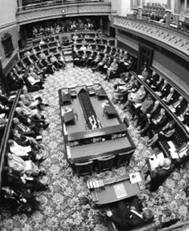 The legislative assembly of the New South Wales Parliament is in session, with members sitting around the house and the speaker sitting at one end. A man in front of a microphone is standing to the right of the speaker.