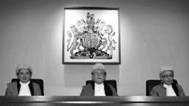 A male judge sits between two female judges at a bench in front of the Australian coat of arms. All three judges are wearing white wigs.