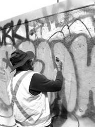 A man, seen from behind holding a hose, stands in front of a concrete wall covered in graffiti.