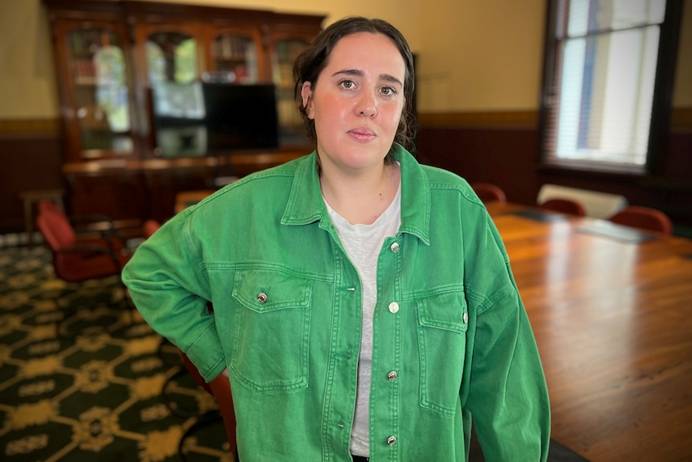 Clare Elliott, in a green bomber jacket & white T-shirt, stands with her right arm resting on her right hip in a heritage room.