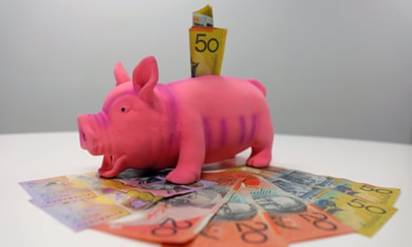 A stock image of a bright pink piggy bank with Australian notes sticking out of it and also on the table beneath it