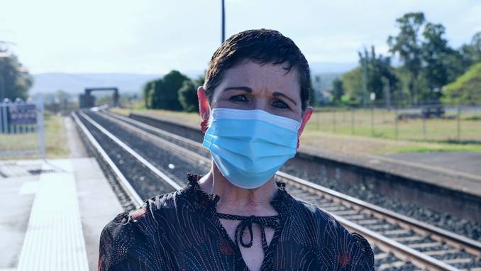 A masked woman with dark hair and clothes stands on a rail line with her arms folded.